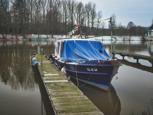 Zeigt ein Boot im Yachthafen. Es is Blau und liegt an einem schlanken verwitterten Steg.